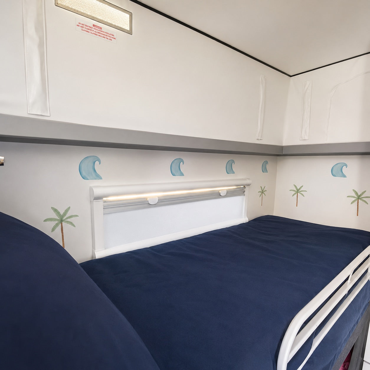 A twin bunk bed with navy blue bedding in the top bunk, featuring palm tree and wave decals on the wall.