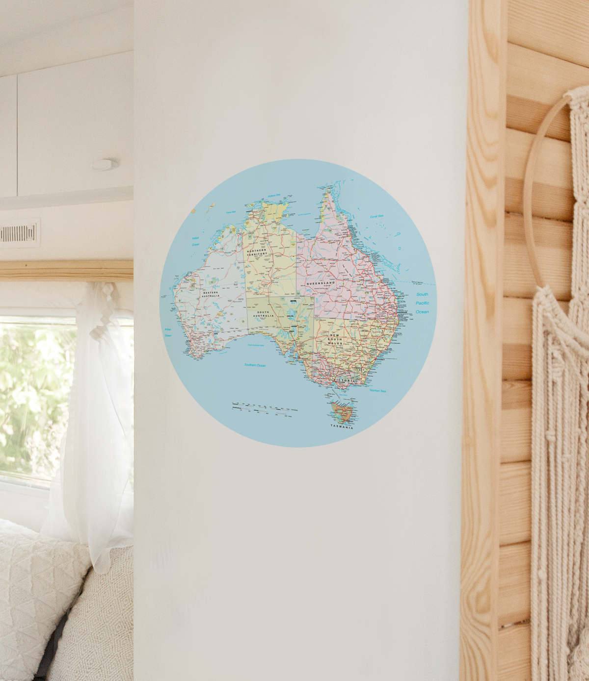 A circular wall map of Australia in pastel colours, displayed on a white wall in a bright, modern room with light wood paneling and a macrame wall hanging.