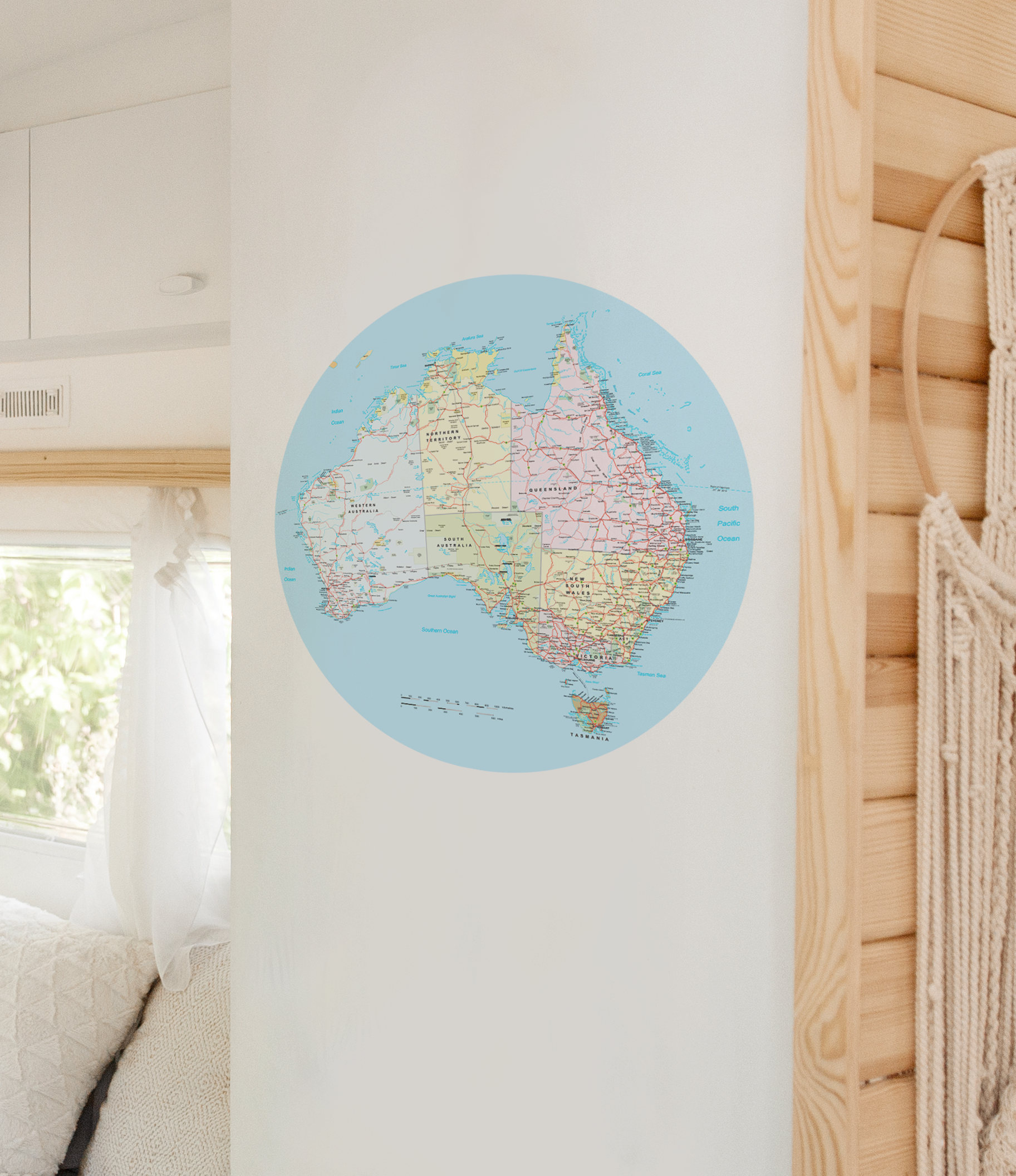 A circular wall map of Australia in pastel colours, displayed on a white wall in a bright, modern room with light wood paneling and a macrame wall hanging.